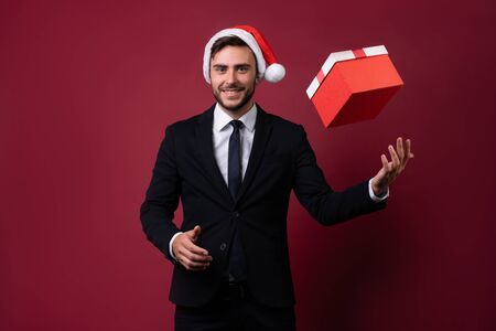 Young handsome caucasian guy in business suit and Santa hats stands on white background in studio and smilie throws up red gift box in hand. Portrait business person with Christmas mood Holiday bannerの写真素材