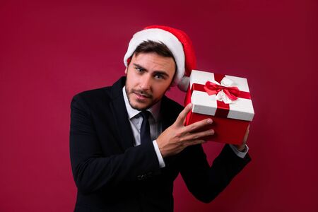 Young handsome caucasian guy in business suit and Santa hats stands on white background in studio and smilie Holds near the ear red gift box Portrait business person with Christmas mood Holiday bannerの写真素材