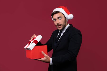 Surprised handsome caucasian guy in business suit and Santa hats stands on white background in studio and smilie opens red gift box in hand. Portrait business person with Christmas mood Holiday bannerの写真素材