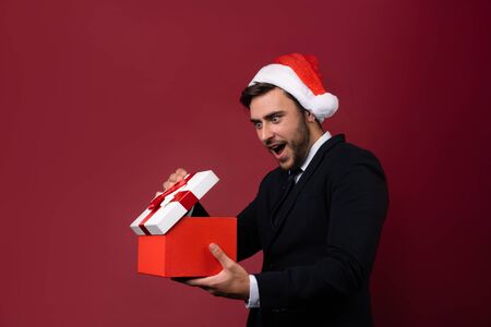 Surprised handsome caucasian guy in business suit and Santa hats stands on white background in studio and smilie opens red gift box in hand. Portrait business person with Christmas mood Holiday bannerの写真素材