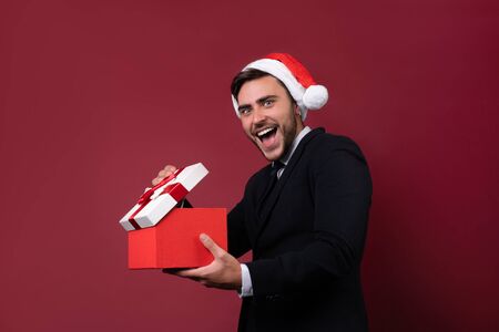 Surprised handsome caucasian guy in business suit and Santa hats stands on white background in studio and smilie opens red gift box in hand. Portrait business person with Christmas mood Holiday bannerの写真素材