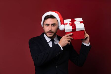 Young handsome caucasian guy in business suit and Santa hats stands on white background in studio and smilie Holds near the ear red gift box Portrait business person with Christmas mood Holiday bannerの写真素材