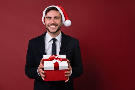 Young handsome caucasian guy in business suit and Santa hats stands on white background in studio and smilie Holding red gift box in hand. Portrait business person with Christmas mood Holiday bannerの写真素材