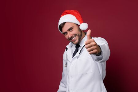 Young handsome doctor in white uniforme and Santa Claus hat standing in studio on red background smile and showing a thumbs up sign Portrait medical student Christmas New Year Holiday Medicine conceptの写真素材