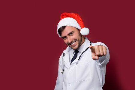 Young handsome doctor in white uniforme and Santa Claus hat standing in studio on red background smile and showing finger in camera Portrait medical student Christmas New Year Holiday Medicine conceptの写真素材