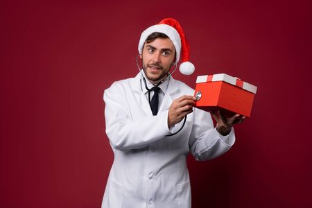 Young handsome doctor in white uniforme and Santa Claus hat standing in studio on red background smile Holding red gift box in hand Portrait medical student Christmas New Year Holiday Medicine conceptの写真素材