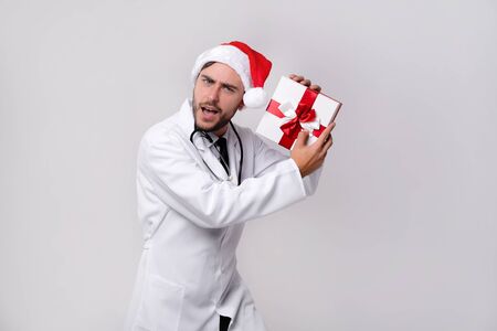 Young handsome doctor in white uniforme  Santa Claus hat standing in studio on white background smile Holding red gift box in hand near ear Medical student Christmas New Year Holiday Medicine conceptの写真素材
