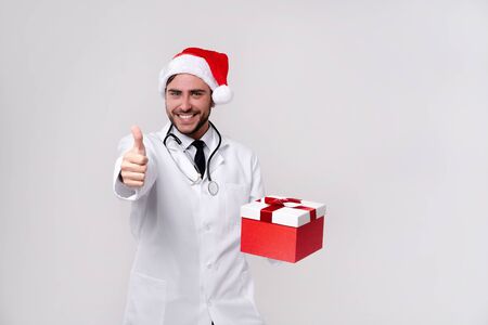 Young handsome doctor in white uniforme and Santa hat in studio white background smile Holding red gift box in hand showing thumbs up Medical student Christmas New Year Holiday Medicine conceptの写真素材