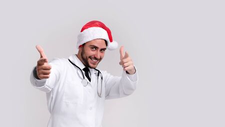 Young handsome doctor in white uniforme and Santa Claus hat standing in studio on white background smile and showing finger in camera Medical student Christmas New Year Holiday Medicine conceptの写真素材