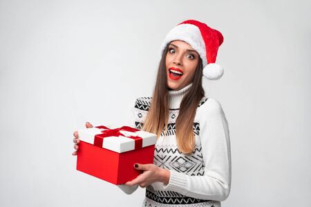 Close up portrait beautifiul caucasian woman in red Santa hat on white studio background. Christmas New Year holiday concept Surprised Cute girl teeth smiling positive emotions with red gift boxの写真素材