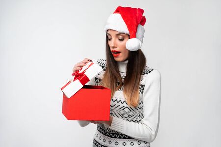 Close up portrait beautiful caucasian woman in red Santa hat on white studio background. Christmas New Year holiday concept Surprised Cute girl teeth smiling positive emotions open big red gift boxの写真素材
