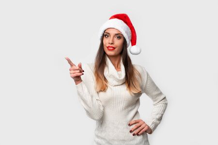 Close up portrait beautiful caucasian woman in red Santa hat on white studio background. Christmas New Year holiday concept. Cute girl teeth smiling positive Showing forefinger finger sideの写真素材