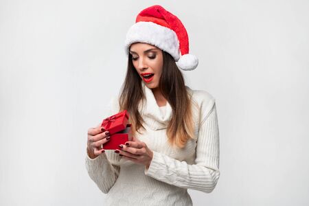 Close up portrait beautifiul caucasian woman in red Santa hat on white studio background. Christmas New Year holiday concept Surprised Cute girl smiling positive emotions open small red gift boxの写真素材