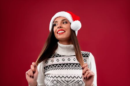 Close up portrait beautifiul caucasian woman in red Santa hat on red studio background. Christmas New Year holiday concept. Cute girl teeth smiling positive emotions touches hair with free copy spaceの写真素材
