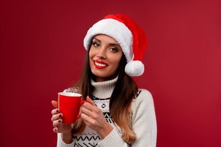Close up portrait beautifiul caucasian woman in red Santa hat on red studio background. Christmas New Year holiday concept. Cute girl teeth smiling positive emotions with Red cup of coffee in handsの写真素材
