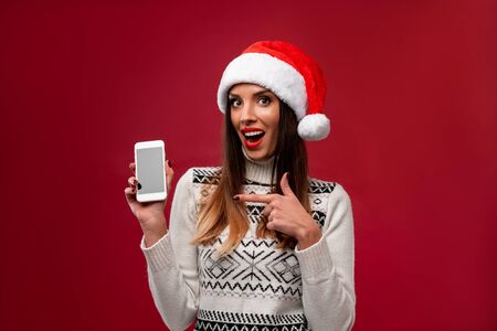 Close up portrait beautiful caucasian woman in red Santa hat on red studio background. Christmas New Year holiday concept. Cute girl teeth smiling positive emotions with free copy spaceの写真素材