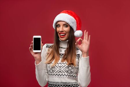 Close up portrait beautiful caucasian woman in red Santa hat on red studio background. Christmas New Year holiday concept. Cute girl teeth smiling positive emotions with free copy spaceの写真素材