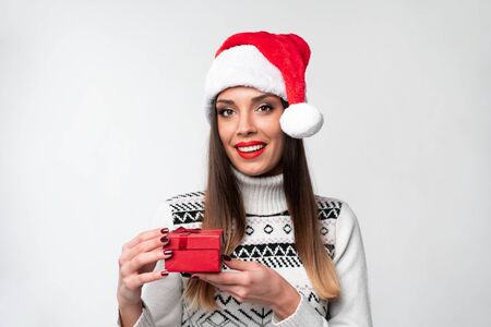 Close up portrait beautiful caucasian woman in red Santa hat on white studio background. Christmas New Year holiday concept Surprised Cute girl teeth smiling positive emotions with small red gift boxの写真素材