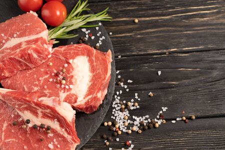 Three pieces of juicy raw beef with rosemary on a stone cutting board on a black wooden table background. Meat for barbecue or grill next to cherry tomatoes sprinkled with pepper and salt seasoningの写真素材