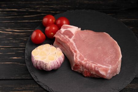 Top view of one pieces raw pork chop steaks with cherry tomatoes and garlic on a black stone cutting board.. Wooden table.の写真素材
