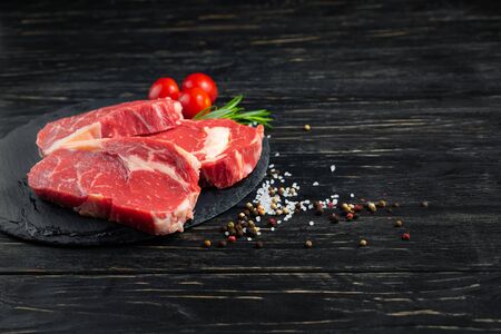 Three pieces of juicy raw beef with rosemary on a stone cutting board on a black wooden table background. Meat for barbecue or grill next to cherry tomatoes sprinkled with pepper and salt seasoningの写真素材