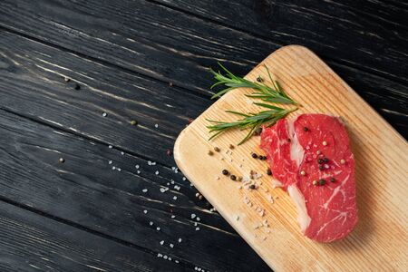 one pieces of juicy raw beef with rosemary on a cutting board on a black wooden table background. Meat for barbecue or grill sprinkled with pepper and salt seasoningの写真素材