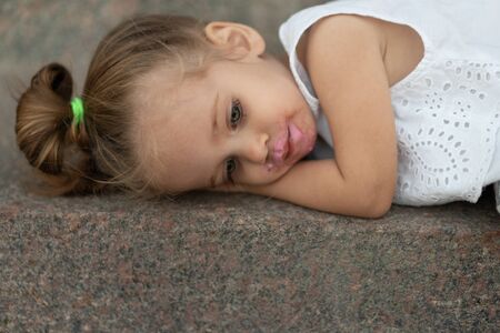 A beautiful little girl of European appearance blonde hair lying sad on street asphalt. Children's problems.Child tired. whimsの写真素材