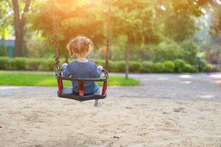 Unrecognizable Little Caucasian girl riding swing at playground sunny summer day rear view. European child having fun outdoor. Family leisure on park. Funny child enjoy the summertime.の写真素材