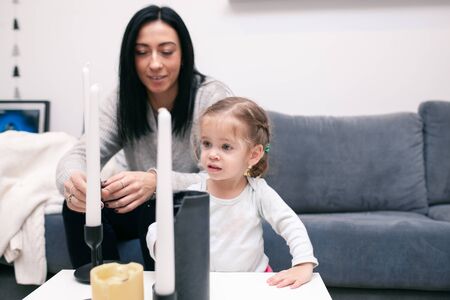 Little girl stands near the sofa at home, pulls his hands to the candlestick, looking into the camera. On the side, her mother sits and watches the girl.の写真素材