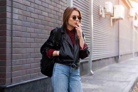 Beautiful young stylishly dressed Caucasian girl smokes cigarette on street Smoking bad habit Nicotine addiction Unhealthy lifestyle Modern woman leather jacket and jeans walking outdoor sunglassesの写真素材