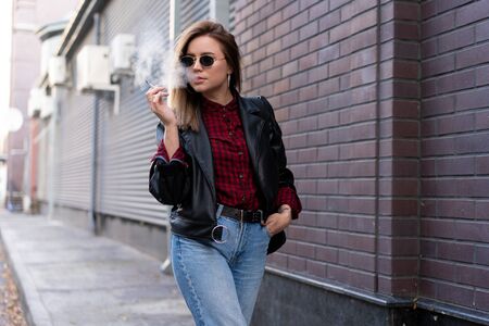 Beautiful young stylishly dressed Caucasian girl smokes cigarette on street Smoking bad habit Nicotine addiction Unhealthy lifestyle Modern woman leather jacket and jeans walking outdoor sunglassesの写真素材