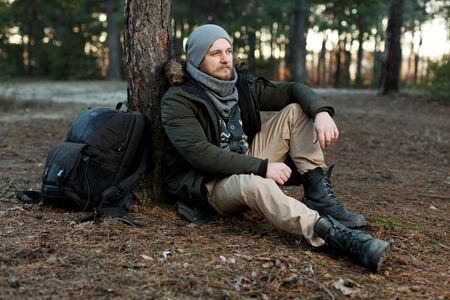 Pensive hipster guy wanderlust with touristic backpack enjoy tranquility during exploring nature environment, male hiker thinking about route to discover sitting on the groundの写真素材