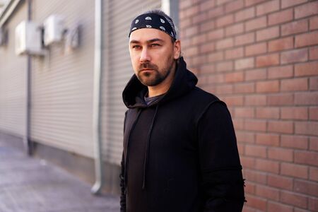 Handsome young unshaven man black hoodie and bandana stands backyard near brick walls summer day. Bearded hipster standing outdoor. Fashionable young adult caucasian guy modern urban clothesの写真素材