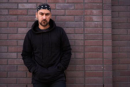 Handsome young unshaven man black hoodie and bandana stands backyard near brick walls summer day. Bearded hipster standing outdoor. Fashionable young adult caucasian guy modern urban clothesの写真素材