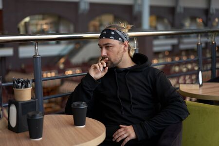 Young handsome lonely man sits cafe table sad waiting for meeting drinking coffee from a paper cup. Caucasian stylish guy dressed black hoodie bandana feel lonely.の写真素材