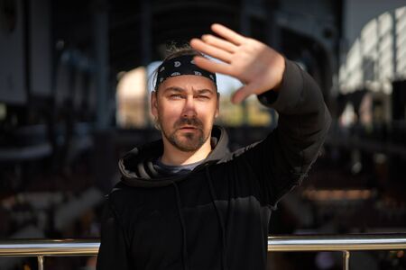 Handsome young unshaven man black hoodie and bandana stands. Bearded hipster standing indoor near handrail. Fashionable young adult caucasian guy modern urban clothes A game of shadowsの写真素材