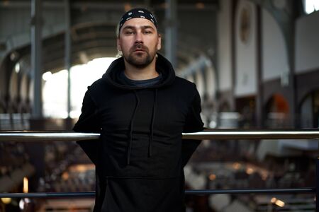Handsome young unshaven man black hoodie and bandana stands. Bearded hipster standing indoor near handrail. Fashionable young adult caucasian guy modern urban clothes A game of shadowsの写真素材