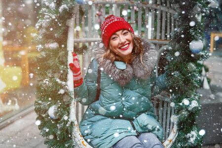 Young beautiful caucasian girl dressed in a warm winter jacket, knitted hat and gloves sits on...