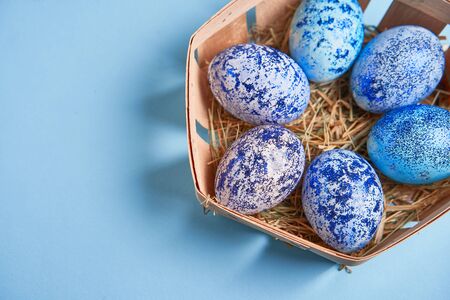 Blue chicken eggs lie in round wooden basket which stands on a blue paper background. Easter...