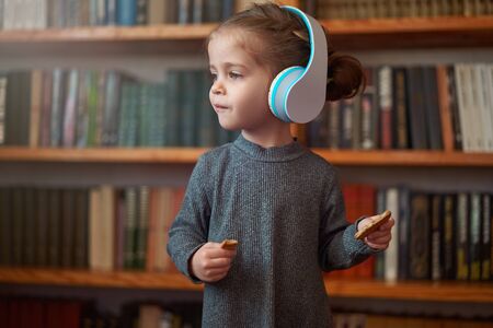 Cute Caucasian little girl in big white headphones listens music in the interior against the background of bookshelves Modern children. Happy childhood with technology Child spends free time at home.の写真素材