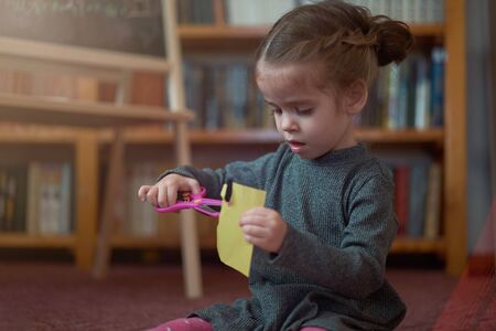 Cute little caucasian girl is playing with scissors and colored paper while sitting on the floor at home. Handcraft for little children. Home leisureの写真素材