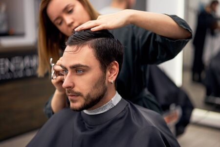 Stylish man sitting barber shop Hairstylist Hairdresser Woman cutting his hair Portrait handsome happy young bearded caucasian guy getting trendy haircut Attractive barber girl working serving client の写真素材