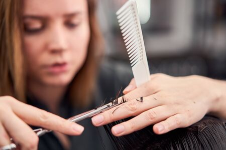 Beautiful caucasian woman hairdresser doing hairstyle to client. Close up portrait female barber with hairdressing tools at work. hair cutting process  Hairstyling in modern barbershop. Professionalの写真素材