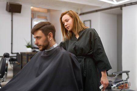 Stylish man sitting barber shop Hairstylist Hairdresser Woman cutting his hair Portrait handsome happy young bearded caucasian guy getting trendy haircut Attractive barber girl working serving client の写真素材