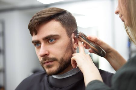 Stylish man sitting barber shop Hairstylist Hairdresser Woman cutting his hair Portrait handsome happy young bearded caucasian guy getting trendy haircut Attractive barber girl working serving client の写真素材
