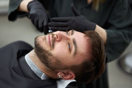 Young adult beautiful caucasian woman hairdresser cuts beard handsome man at modern barbershop Happy gay sitting chair men beauty salon. beard care. metrosexual. Female barber in black gloves servingの写真素材