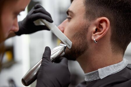 Young adult beautiful caucasian woman hairdresser cuts beard handsome man at modern barbershop Happy gay sitting chair men beauty salon. beard care. metrosexual. Female barber in black gloves servingの写真素材