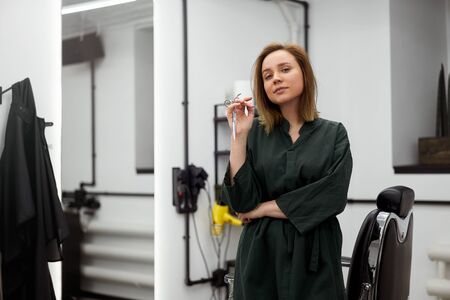 Beautiful caucasian woman hairdresser standing near armchair. Portrait female barber standing near armchair with hairdressing tools  Hairstyling in modern barbershop. Professionalの写真素材