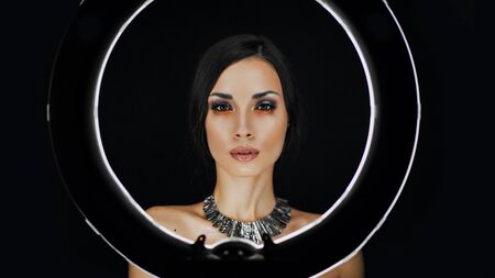 A beautiful young Caucasian girl with a beautiful make-up looks out from behind a circular lamp for...