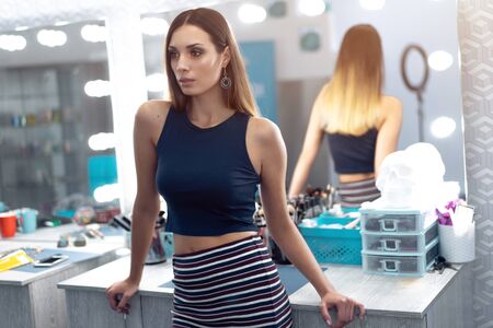 Portrait of pretty female beautician standing posing at workplace in hair and beauty salon with big mirror on the backgroundの写真素材